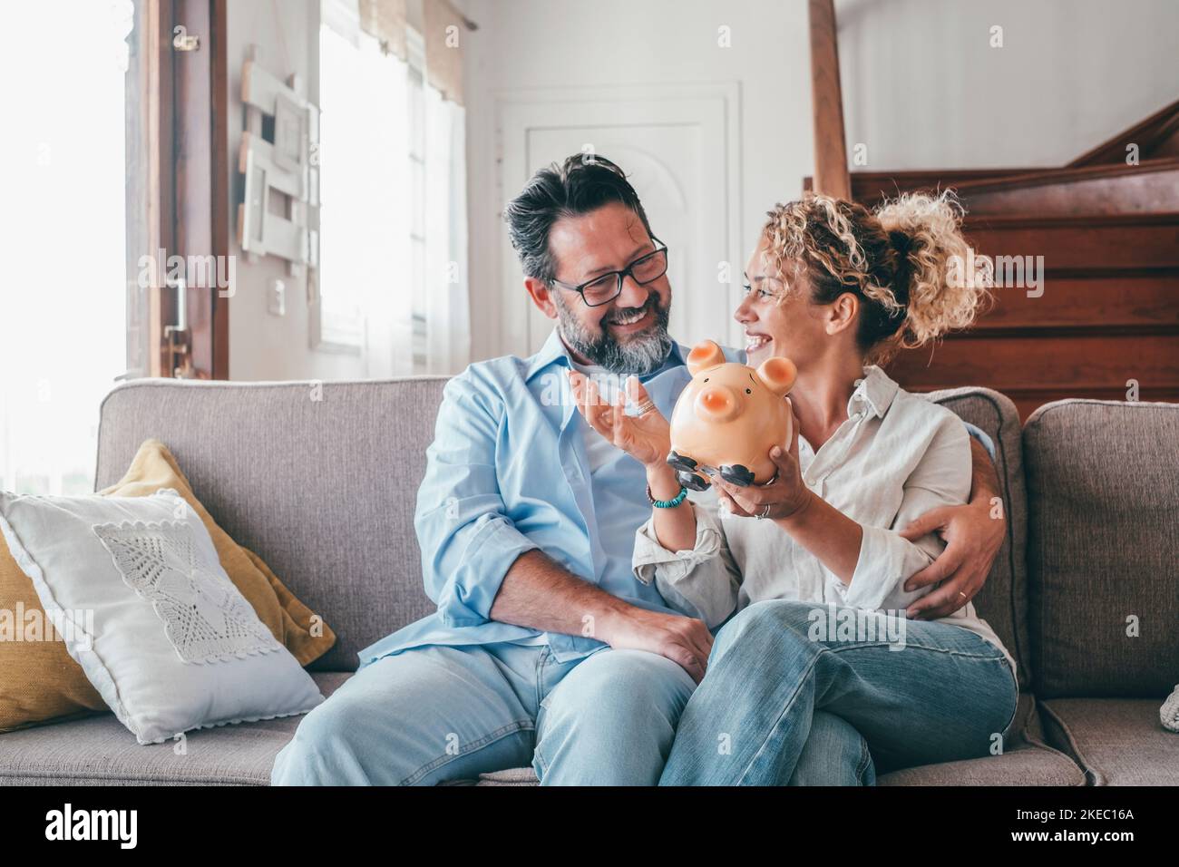 Happy caucasian couple holding piggy bank to save money to make their future dreams come true. Loving man and woman holding piggy bank for savings and discussing while sitting on sofa at home Stock Photo