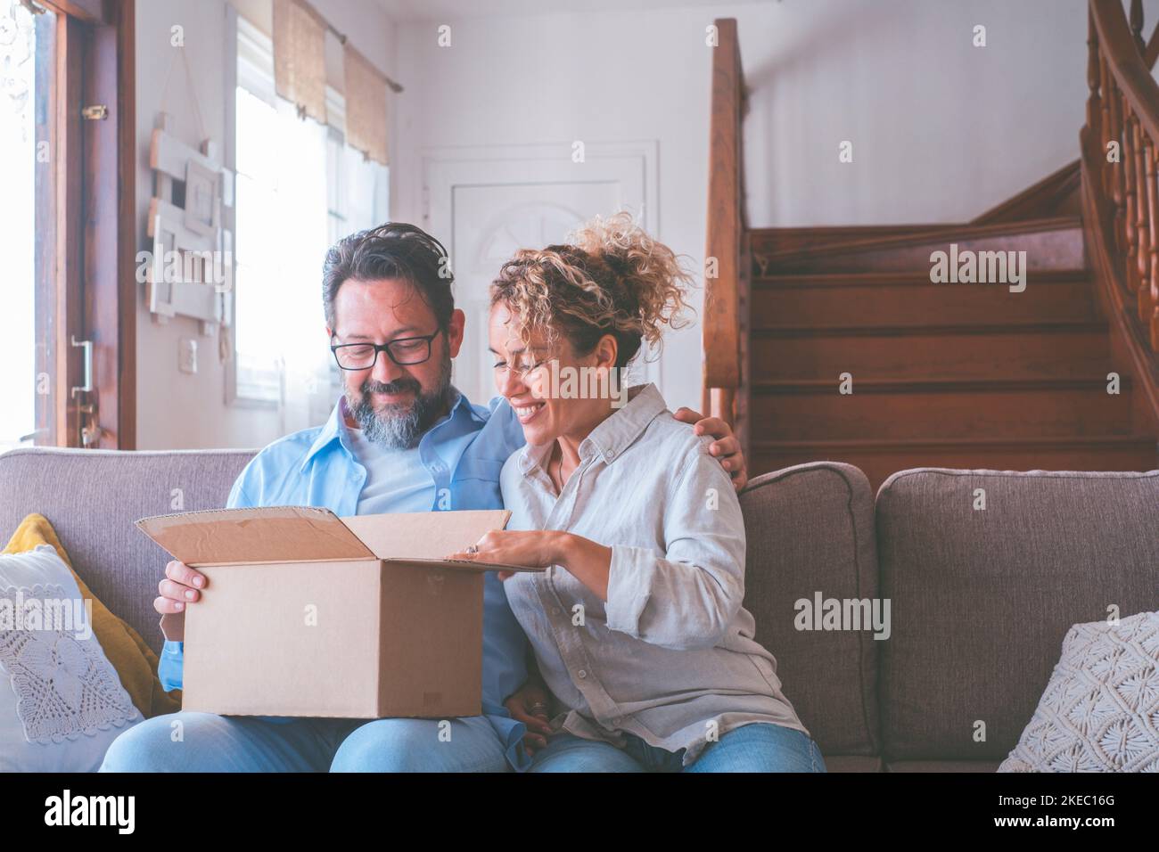 Happy caucasian couple looking inside cardboard box, satisfied with ordered item. Husband and wife sitting on sofa opening carton parcel in living room of house. Man and woman clients unbox package with internet order Stock Photo