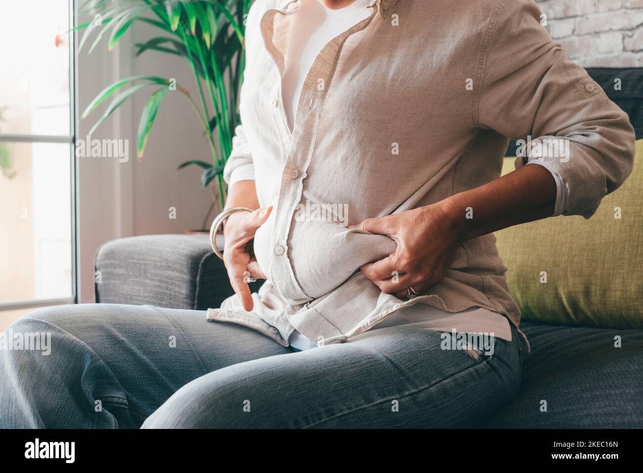 Midsection of woman hands holding her belly fat sitting on sofa at home. Caucasian lady grabbing excessive fat on her abdomen. Young female pinching on her fatty obese waist Stock Photo
