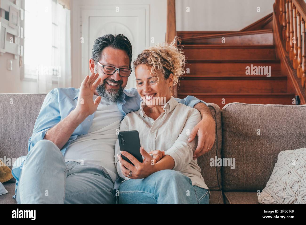 Happy caucasian couple resting on couch, waving hand in hello gesture greeting friends at online video call distant meeting using mobile phone. Wife with husband waving hand while doing video call on smartphone Stock Photo