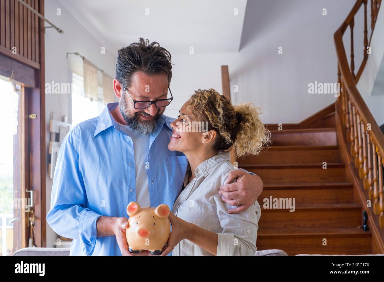 Happy caucasian couple holding piggy bank to save money to make their future dreams come true. Loving man and woman embracing each other while holding piggy bank for savings at home Stock Photo