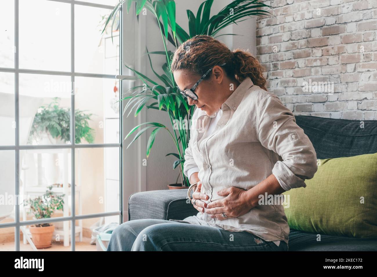 Young woman holding belly suffering from abdomen pain in morning on sofa at home. Caucasian lady touching her stomach in severe ache while sitting on couch in modern living room apartment Stock Photo