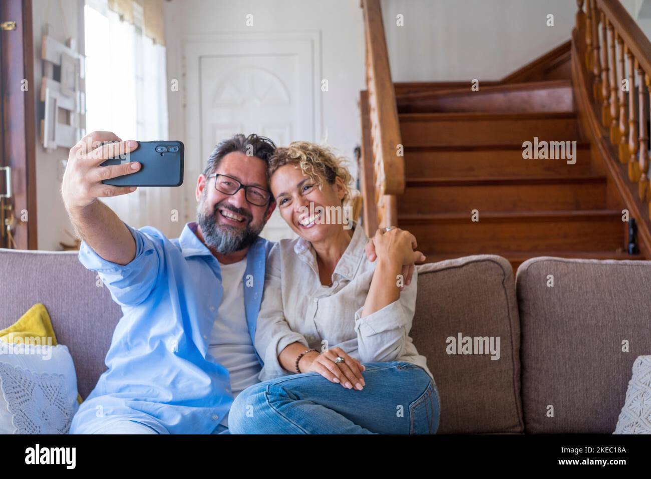 Happy caucasian couple taking selfie sitting on sofa using mobile phone camera at home. Joyful wife and husband taking self portrait on smartphone. Couple enjoying clicking pictures at new apartment Stock Photo