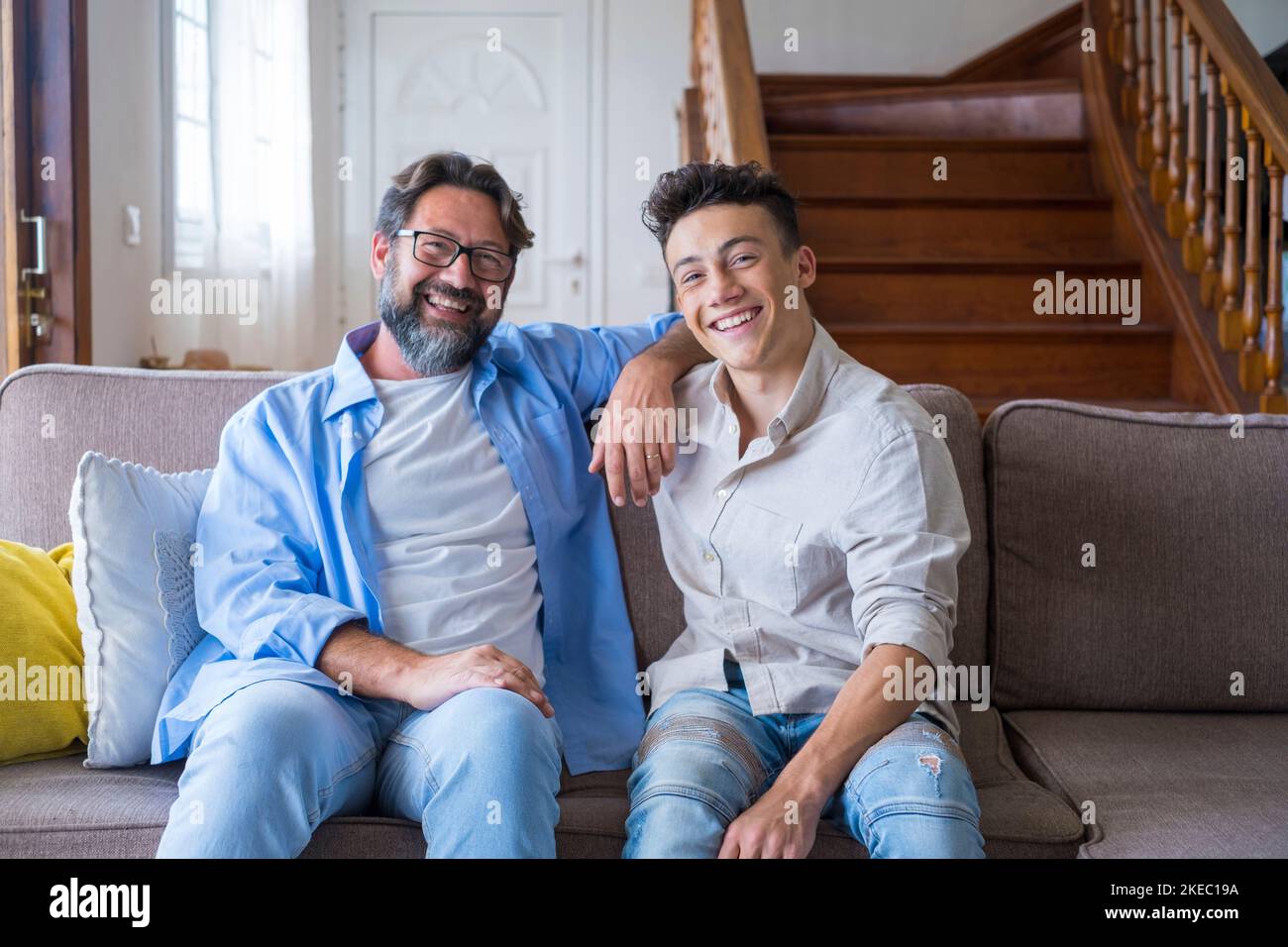 Happy father and son sitting on sofa at home. Cheerful father with teenage son sitting on couch in the living room of apartment. Portrait of father and son spending leisure time at home Stock Photo