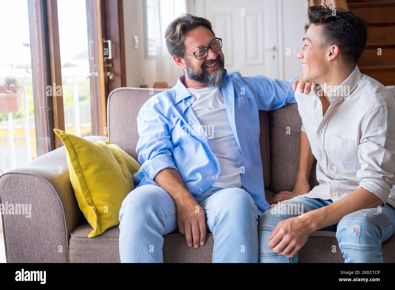 Happy father and son talking while sitting on sofa at home. Cheerful father with teenage son sitting on couch in the living room of apartment. Father and son spending leisure time at home Stock Photo