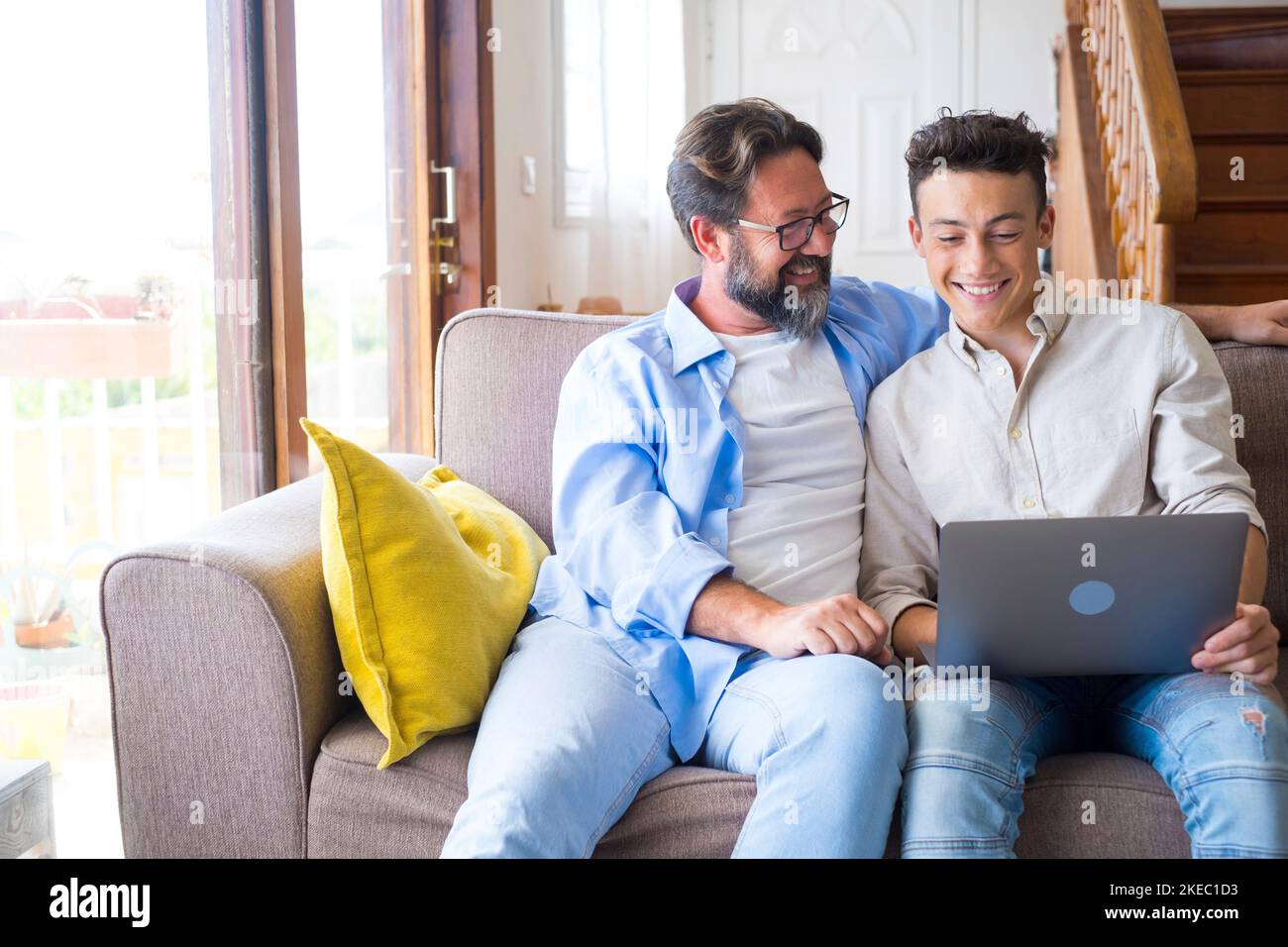 Happy father and son looking at laptop screen while sitting on couch in living room at home. Father with teenage son browsing social media content using laptop in living room Stock Photo