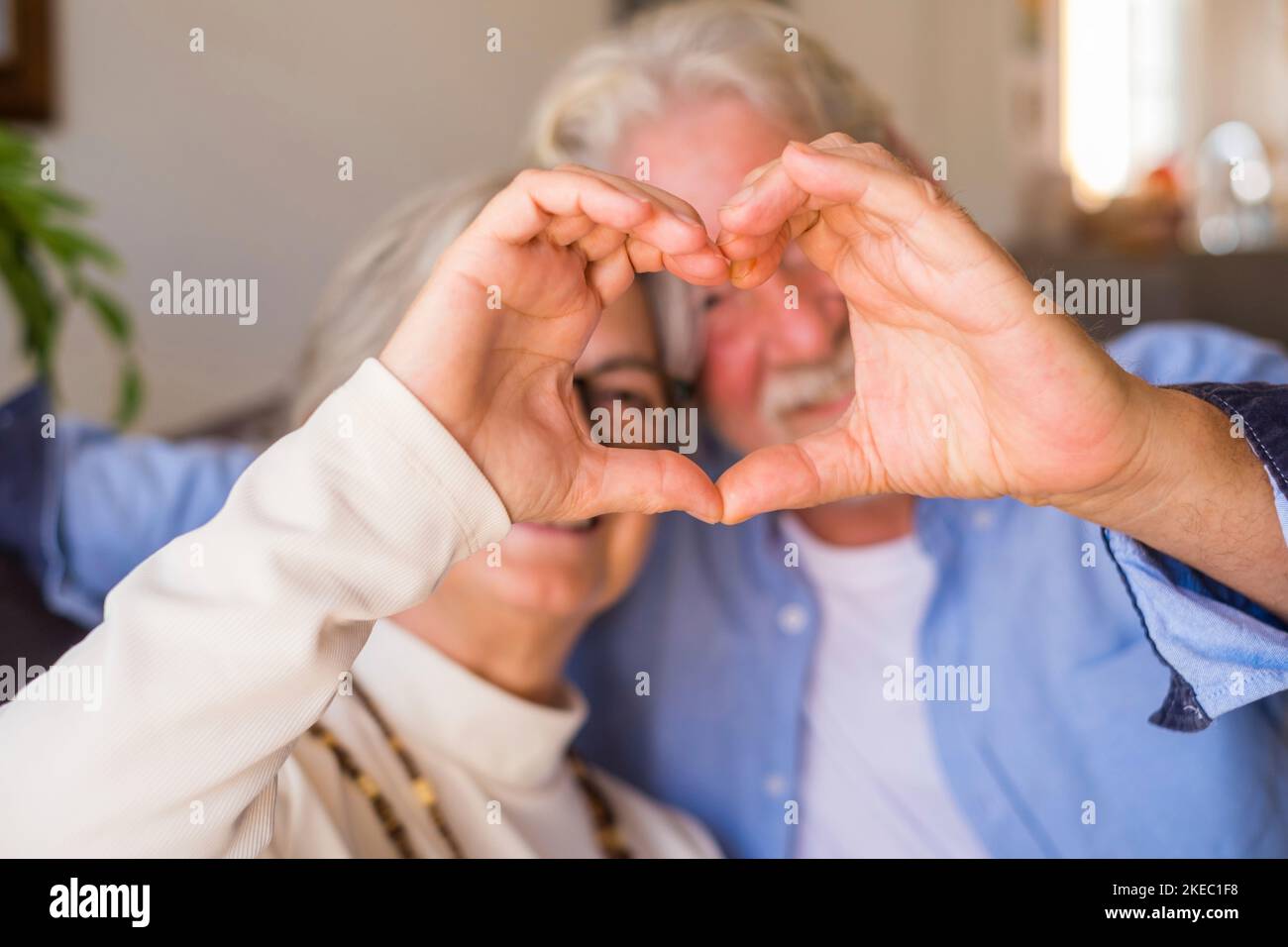 Couple of two old and happy seniors having fun at home on the sofa doing a heart shape with their hands and fingers looking at the camera. Stock Photo