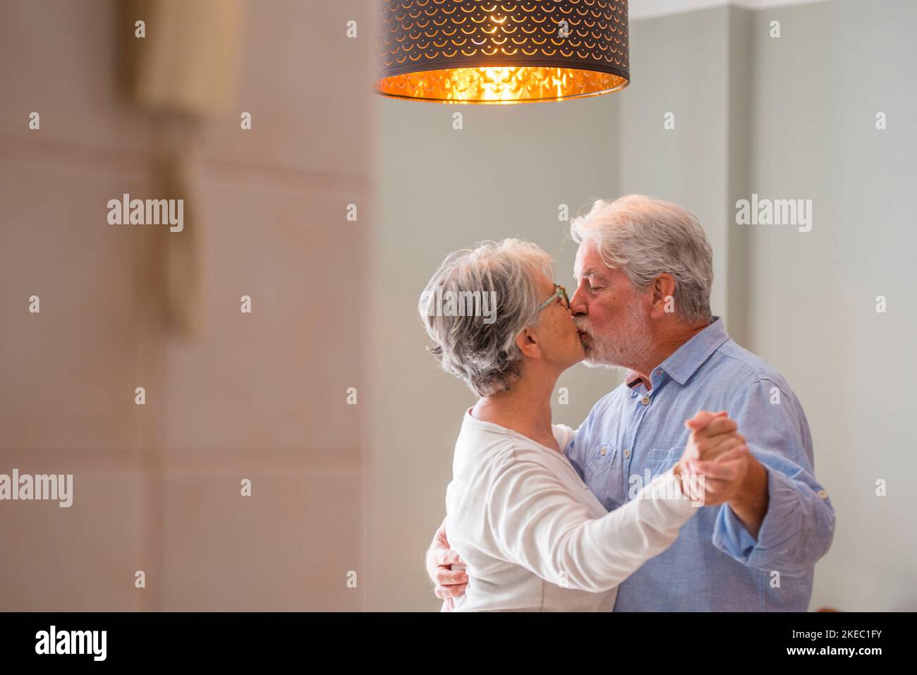 Romantic loving senior couple holding hands while kissing and enjoying dancing together in the living room of house, Elderly happy couple celebrating by doing dance at home. Old husband and wife having fun time at modern apartment Stock Photo