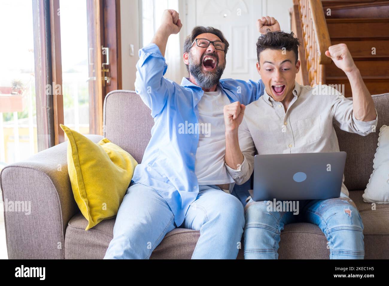 Happy father and son with arms raised celebrating success while sitting with laptop on couch in living room at home. Astonished father with son celebrating online sucess at home Stock Photo
