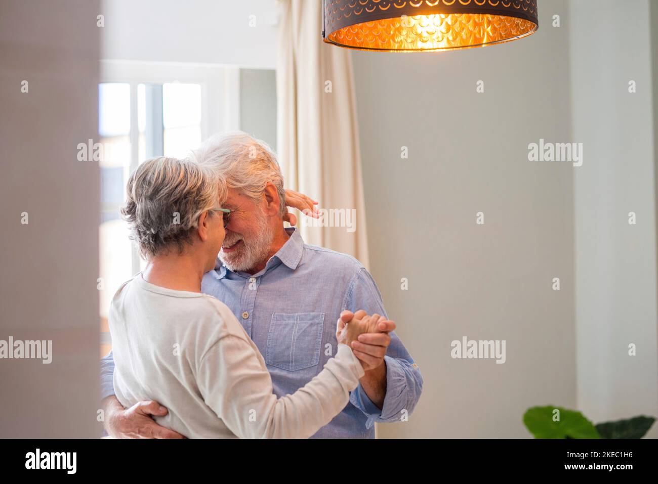 Cheerful senior couple dancing and smiling looking at each other. Elderly happy couple celebrating by dancing in living room. Romantic old aged couple holding hands and dancing together at home Stock Photo