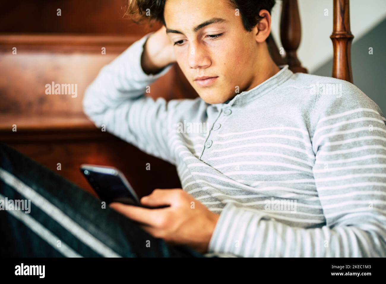 Bored handsome male teeanger at home use cellular phone and internet connection to read new or chat with friends - concept of coronavirus lockdown worldwide quarantine emergency Stock Photo