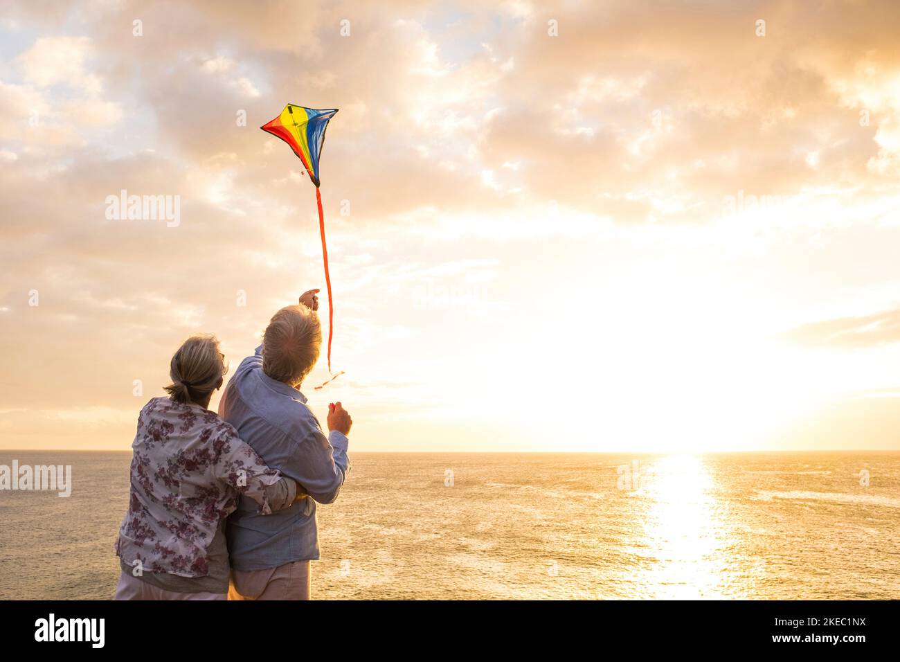 close up and portrait of two old and mature people playing and enjoying with a flaying kite at the beach with the sea at the background with sunset - active seniors having fun Stock Photo