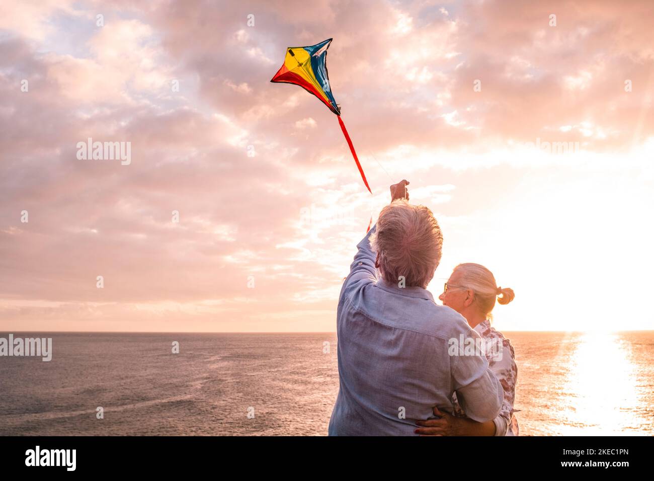 close up and portrait of two old and mature people playing and enjoying with a flaying kite at the beach with the sea at the background with sunset - active seniors having fun Stock Photo