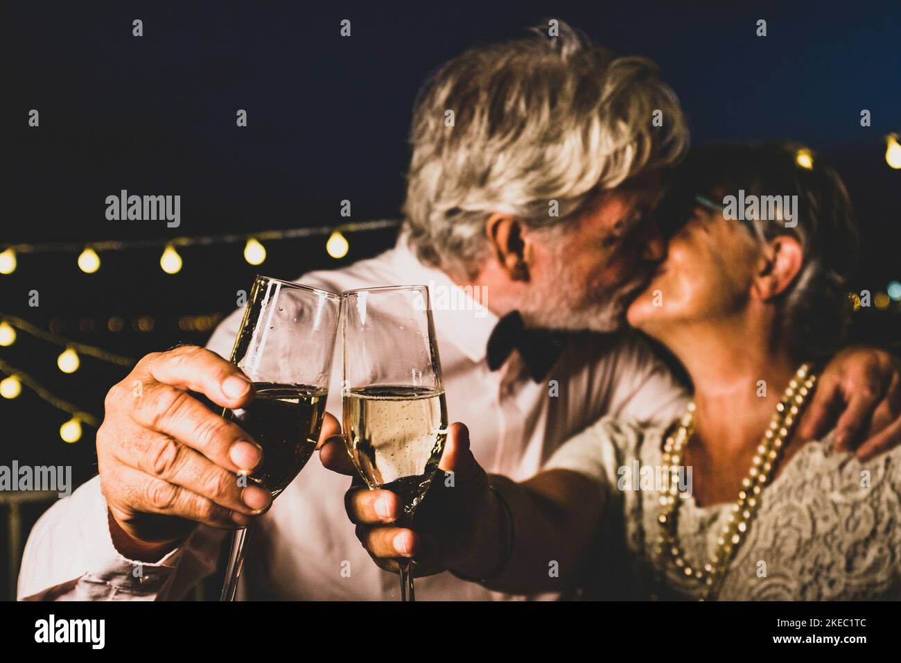 Old caucasian couple kissing each other while toasting champagne glasses celebrating new year party. Loving husband and wife kissing each other while partying with white wine glasses in hands Stock Photo