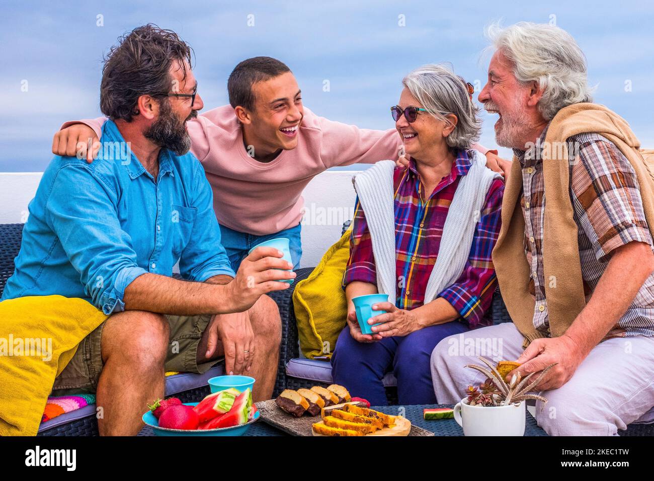group of four people of all ages together at home in the balcony laughng and having fun and eating food like fruit and cookies - teenager enjoying with seniors and middle age man Stock Photo