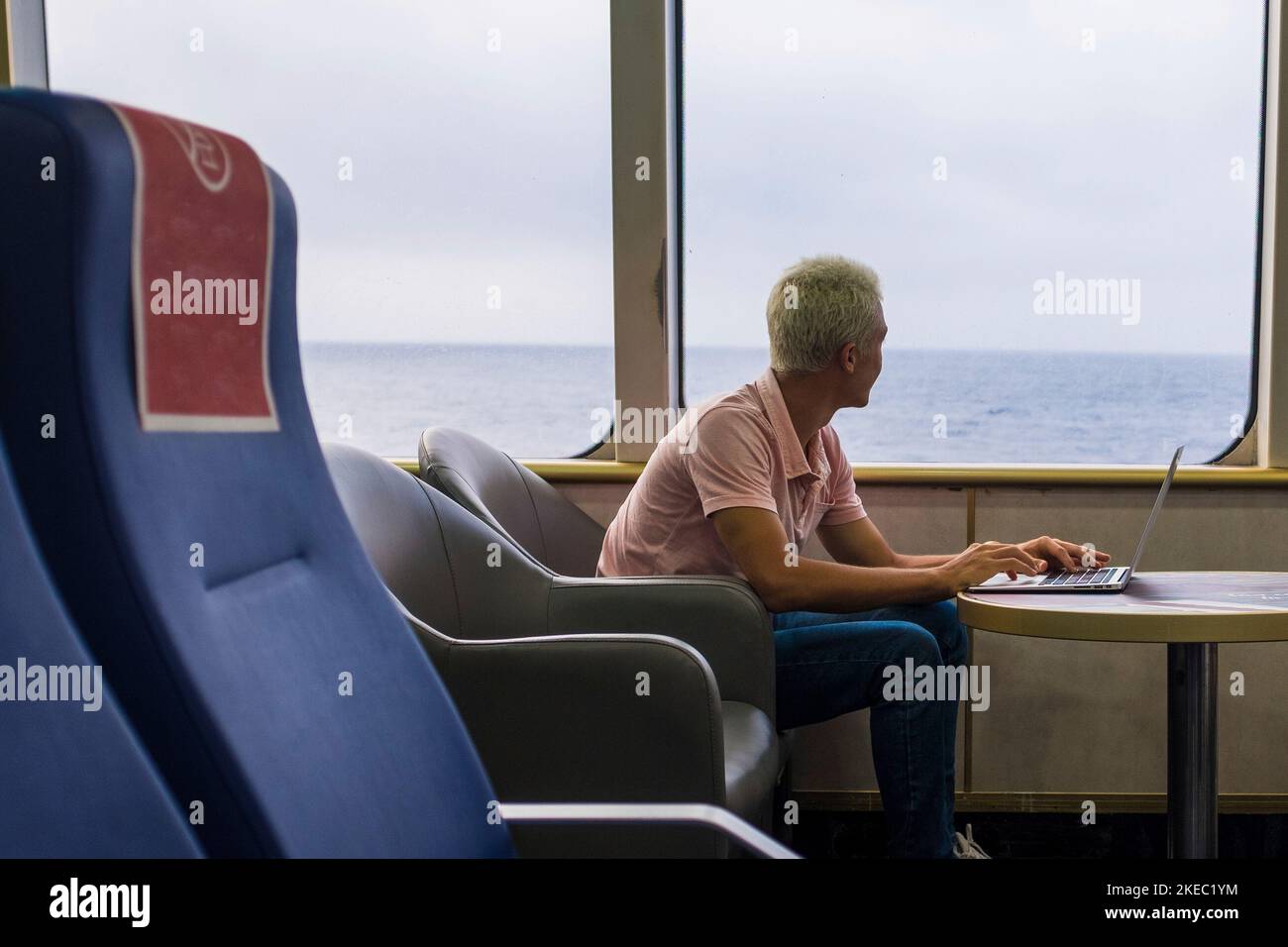 one young man using his laptop or computer pc on a ship traveling in the water of the sea or ocean - man looking outdoor while workig with device - isolated nomad alone people traveling Stock Photo