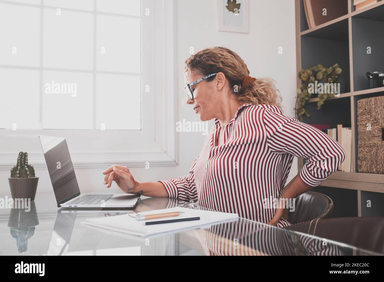 Caucasian woman touching her back for stress pain and disease caused by work posture. Businesswoman suffering with back ache while sitting on office chair and working on laptop. Female executive with lower back pain Stock Photo