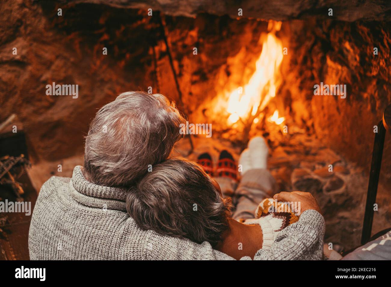 Old caucasian couple spending leisure time together at home. Back of romantic senior husband embracing with wife while relaxing and looking at burning lit fireplace during winter holiday Stock Photo