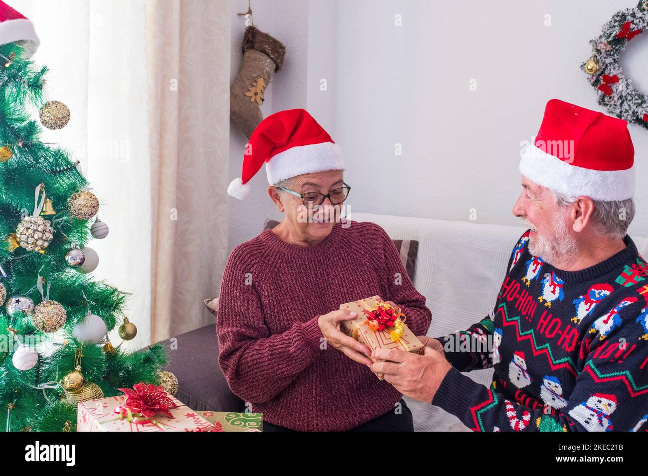 Senior couple in warm clothing and santa hat, man covering eyes of old wife with a surprise gift for her. Loving old romantic heterosexual couple celebrating christmas festival together Stock Photo