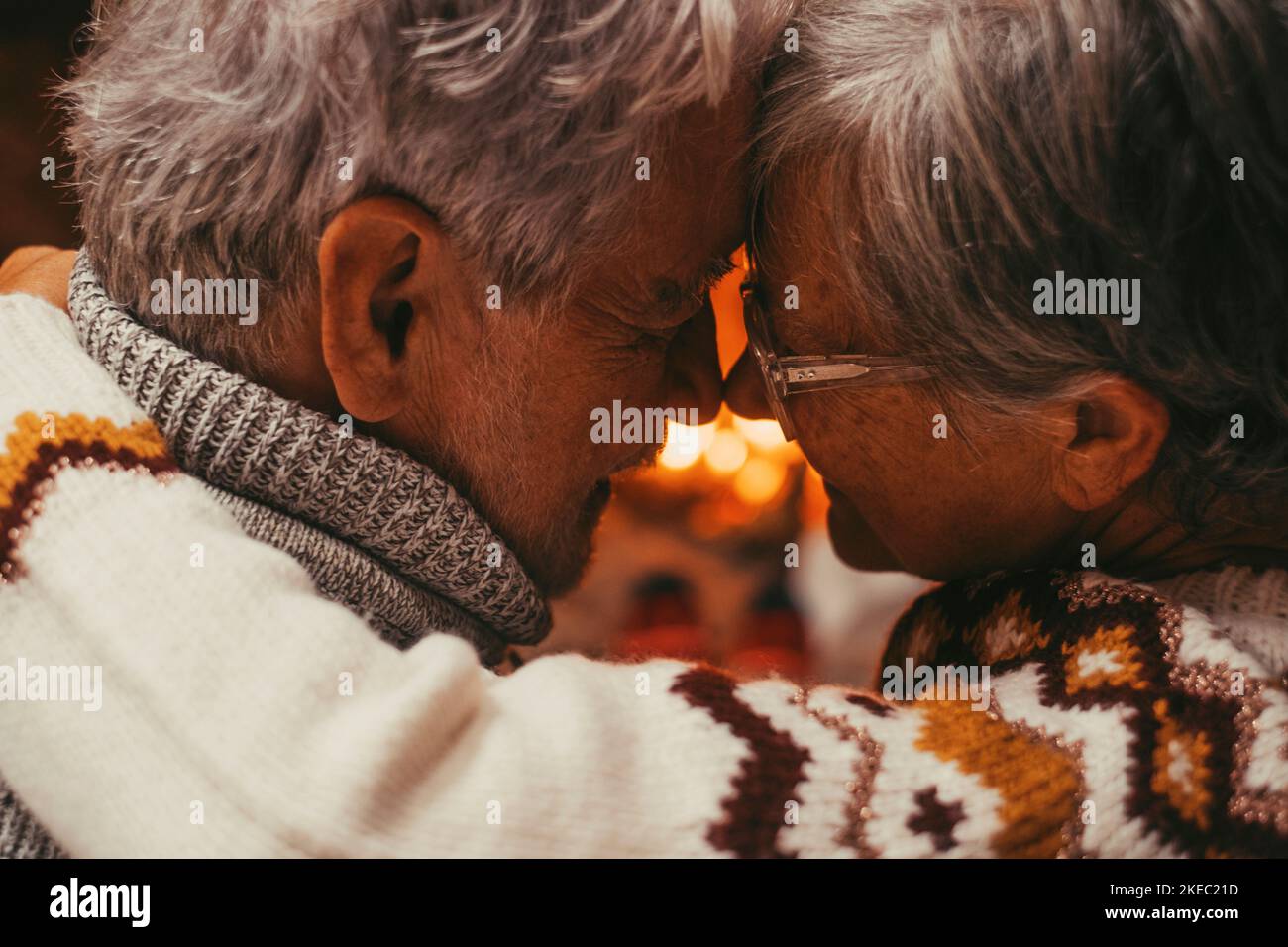 Old caucasian couple spending leisure time together at home. Loving husband and wife touching forehead looking at each other face to face with fireplace in the background during winter holiday Stock Photo