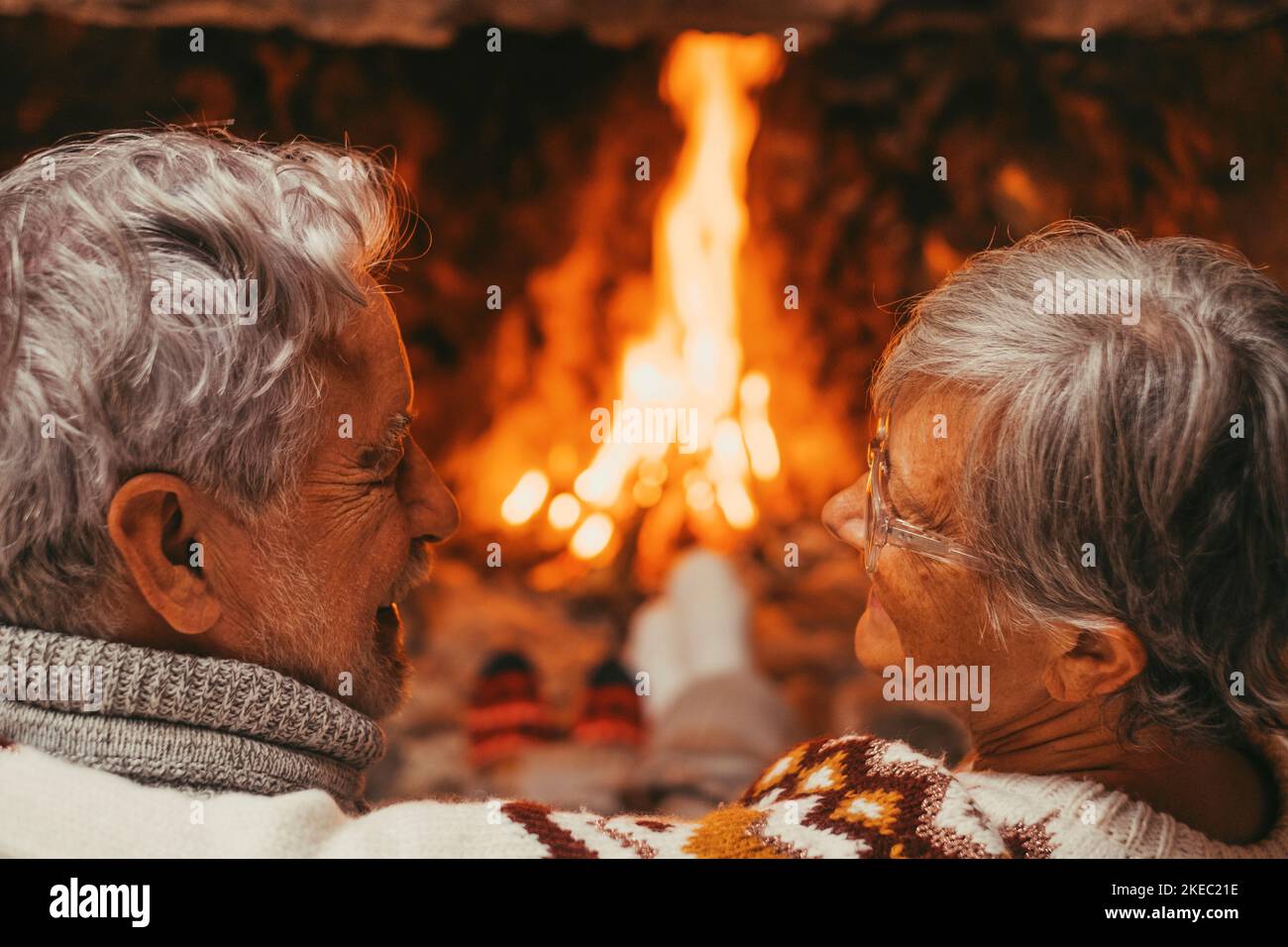 Old caucasian couple spending leisure time together at home. Loving romantic husband and wife looking at each other face to face with fireplace in the background during winter holiday Stock Photo