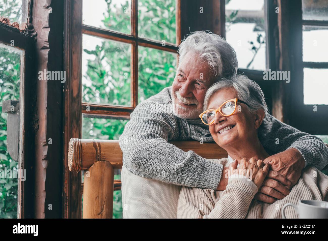 Old caucasian couple spending leisure time looking out through window at home. Loving husband embracing wife from behind sitting on armchair at house and admiring view from transparent door glass. Stock Photo