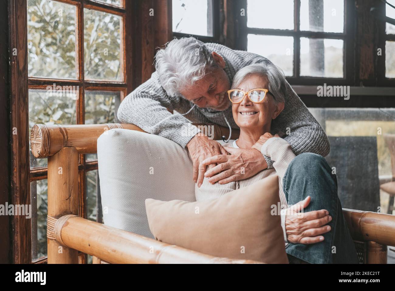 Old caucasian couple spending leisure time together at home. Loving husband embracing wife from behind sitting on armchair at house. Romantic aged man hugging his life partner relaxing on chair Stock Photo