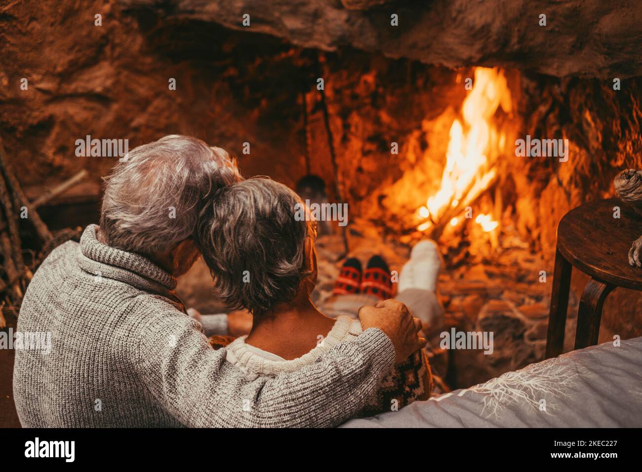 Old caucasian couple spending leisure time together at home. Back of loving romantic husband embracing wife and relaxing while looking at burning lit fireplace during winter holiday Stock Photo