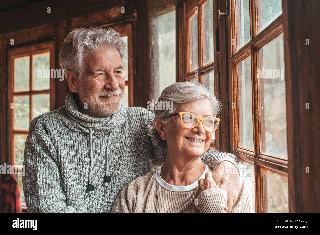Old caucasian couple spending leisure time looking out through window at home. Loving husband embracing wife from behind and admiring something interesting from transparent door glass. Stock Photo