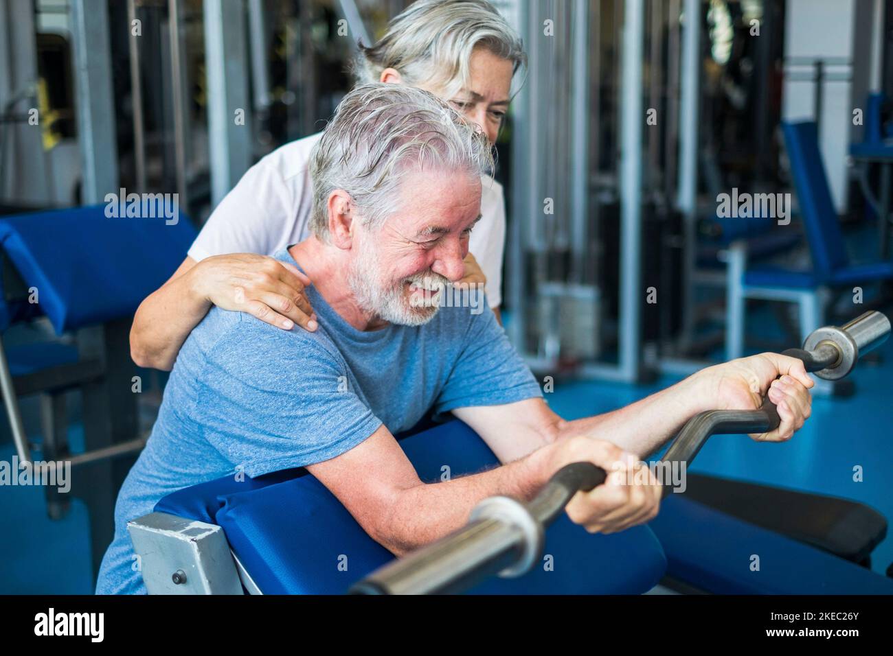 couple of seniors at the gym doing exercise together - mature woman looking and helping her husband holdinng a barr and lifting weight Stock Photo