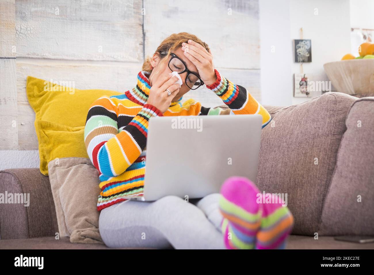 sick and unwell young woman feeling wrong with flu and some virus on the sofa at home. Female with fever and headache using laptop or technology device. Stock Photo