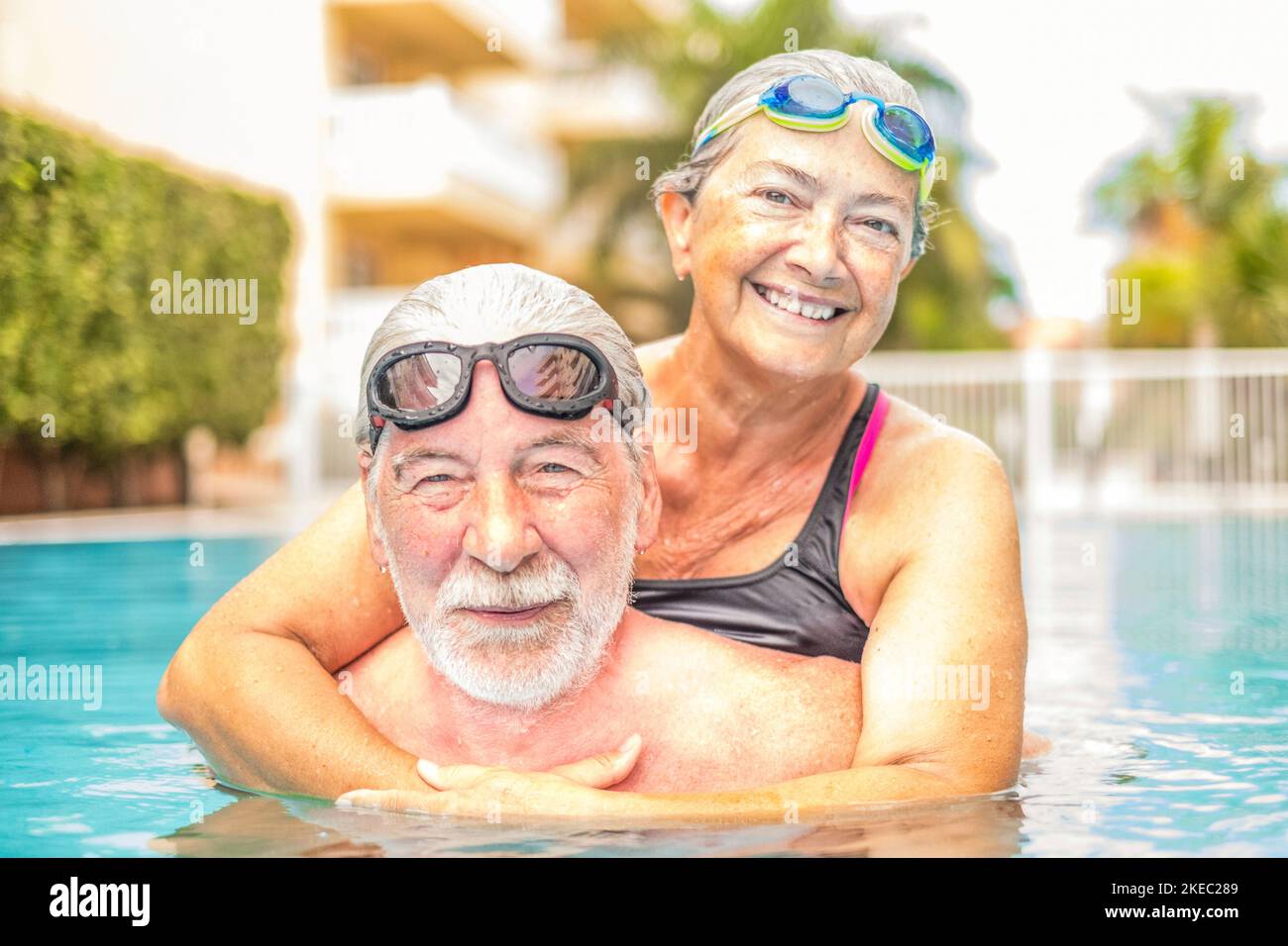 couple of seniors enjoying summer and having fun together in the pool swimming and smiling looking at the camera - cheerful happy people in their vacations - portrait of two mature and retired pesioners Stock Photo