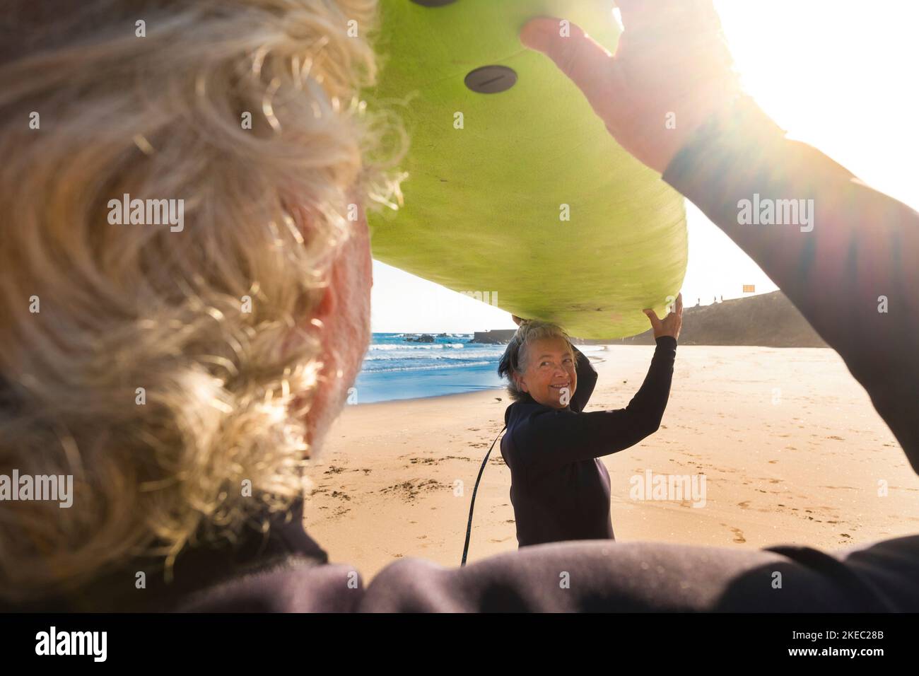 couple of mature and old people or seniors surfing together in the beach with a big surfboard. Two seniors having fun and enjoying summer doing water sports outdoor Stock Photo