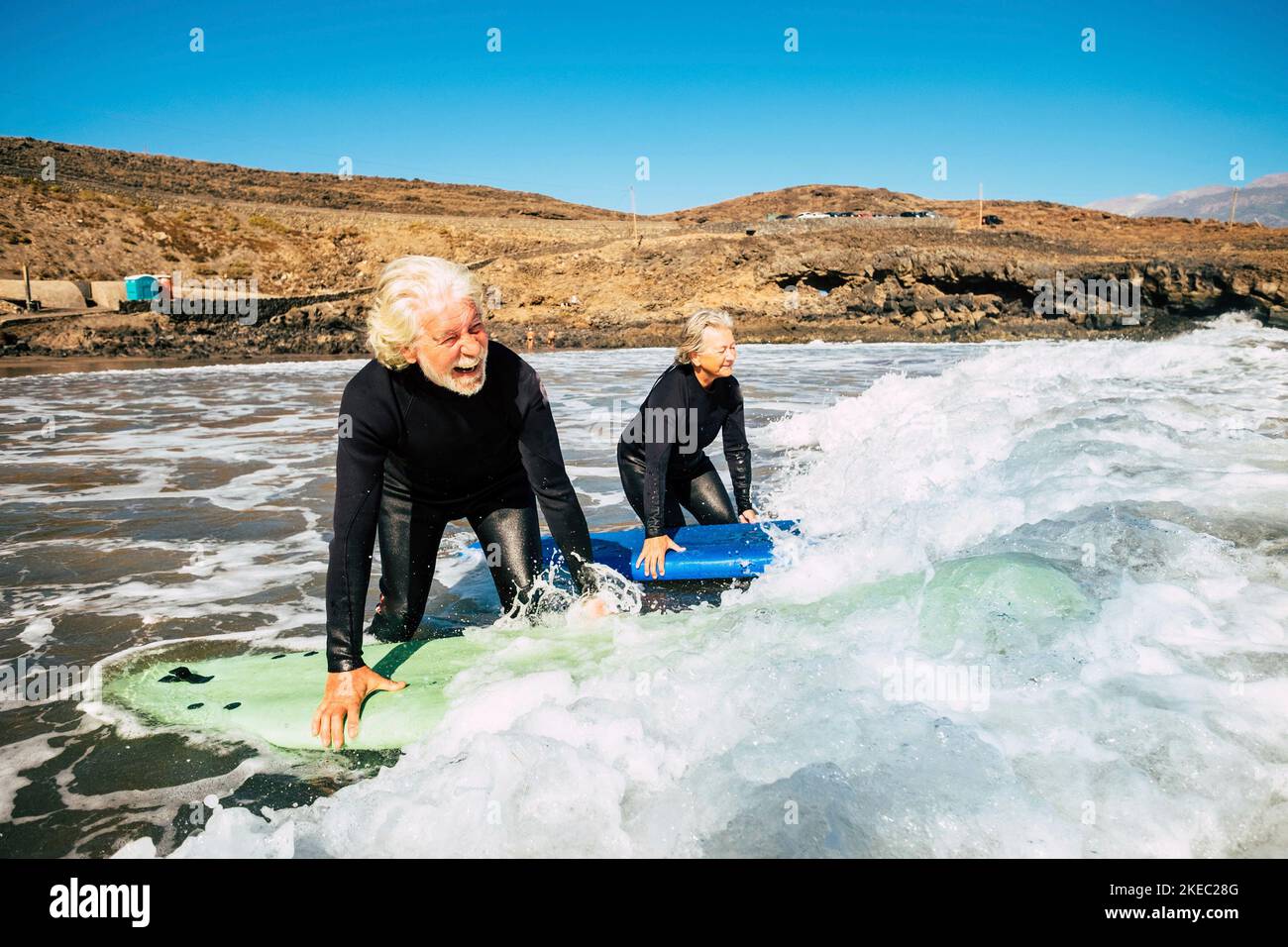 couple of seniors going surfing together - two mature people in the beach with surfboards and wetsuits looking at the sea entring in the water - learning surf Stock Photo