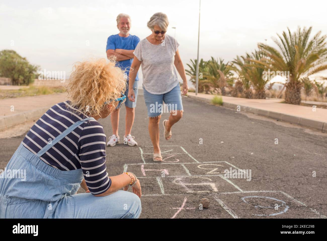 group of three people like adults and senior - two seniors playing at hopscotch with a curly woman looking at the mature woman jumping Stock Photo