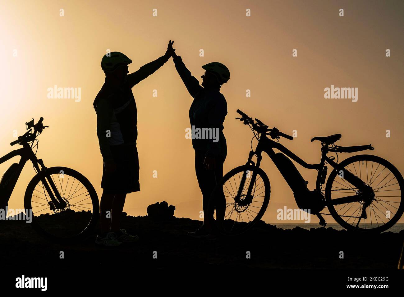 couple of two seniors and mature people together at the sunset with their bikes giving five with the silhouette and the sea at the background Stock Photo