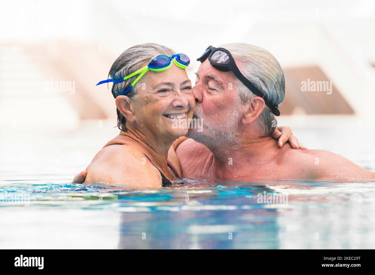 couple of seniors having fun together in a swimming pool smiling - man giving a kiss to his wife Stock Photo