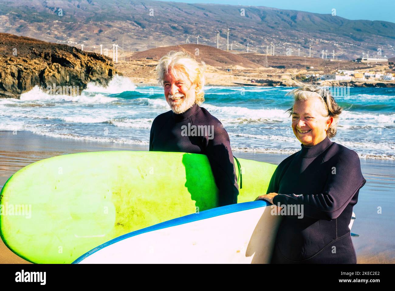 couple of two happy and active seniors at the beach tryin to learn surfing - pensioners with surftables and wetsuits ready to enter in the water and take waves Stock Photo