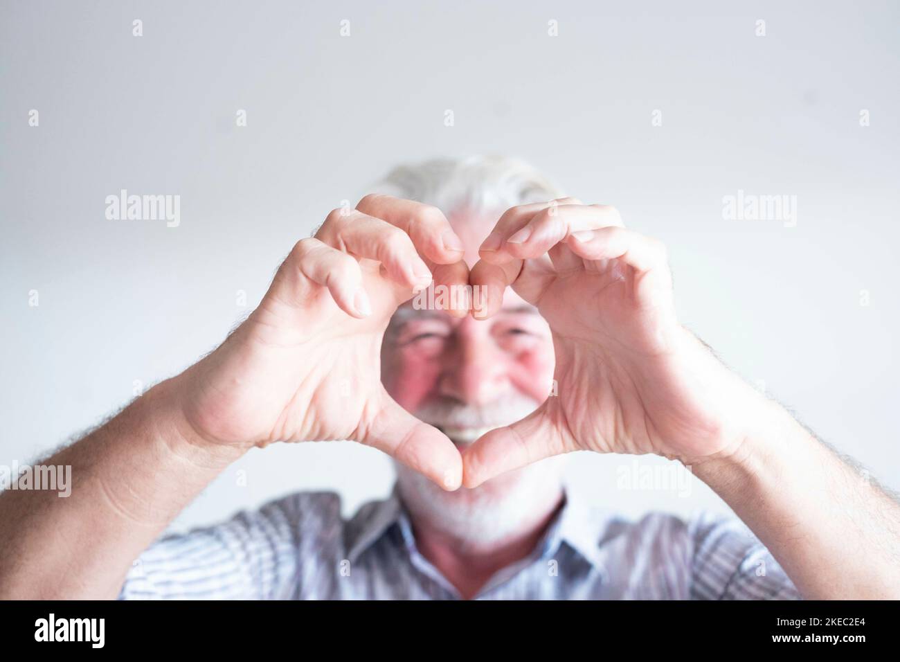 close up and portrait of mature man and senior doing an heart with his fingers and hand in front of the camera - happy pensioner having fun enjoying - white background Stock Photo