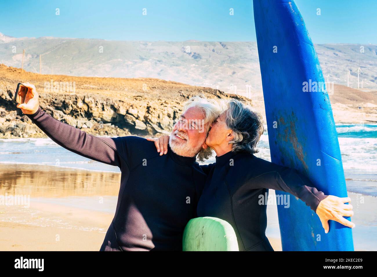 couple of two cute seniors and pensioners doing exercise and taking a selfie with their surftables and wetsuits - woman kissing her husband Stock Photo