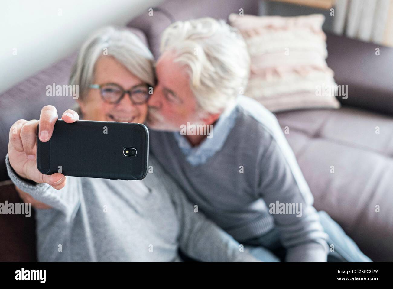 cute couple of two seniors married taking a selfie together sitting on the sofa at home - mature man kissing his wife while she's taking a photo Stock Photo