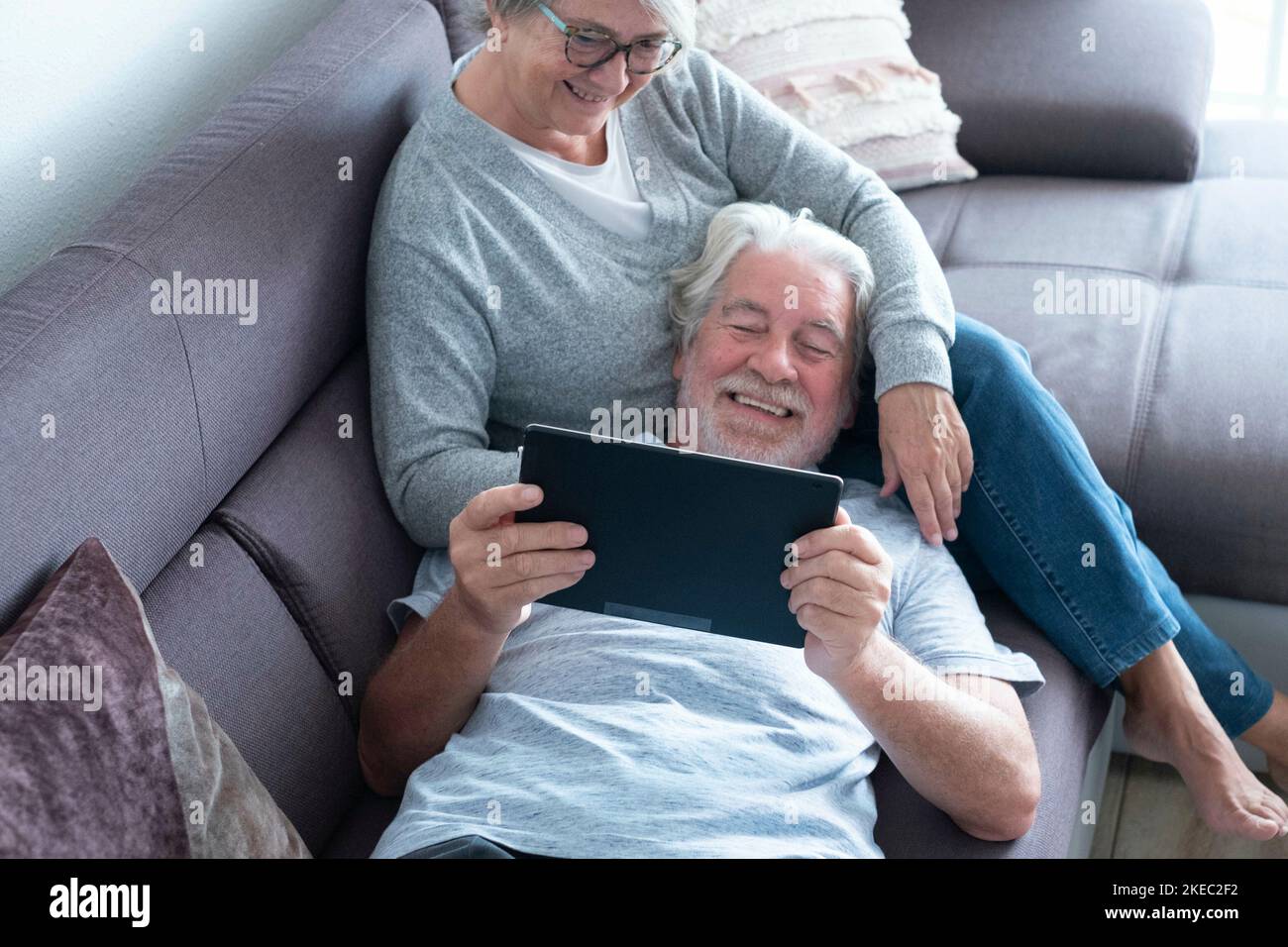 couple of two seniors or mature people at home having fun together looking at the same tablet - pensioner lying on the sofa with his wife sitting Stock Photo