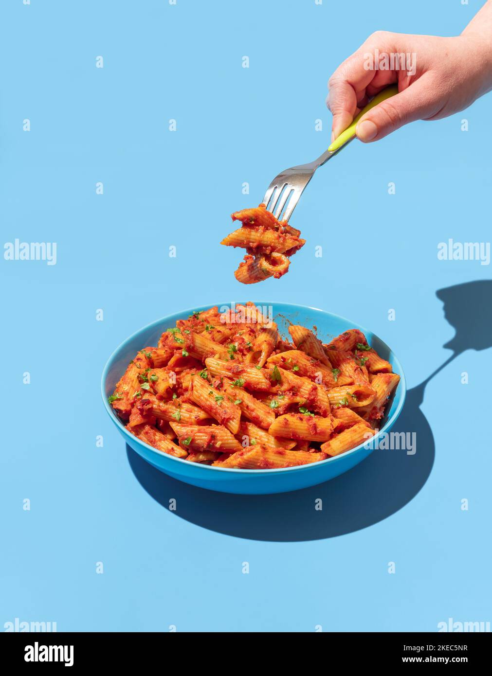 Woman's hand taking pasta with a fork from a blue bowl, minimalist on a blue table. Pasta alla arrabbiata in a blue bowl in bright light. Stock Photo