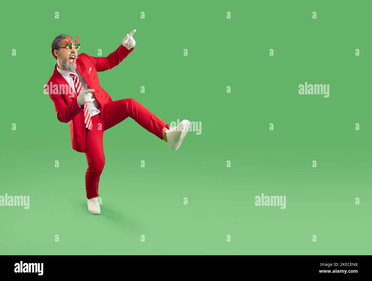 Happy man in red suit and funny Christmas glasses pointing to copy space on green background Stock Photo