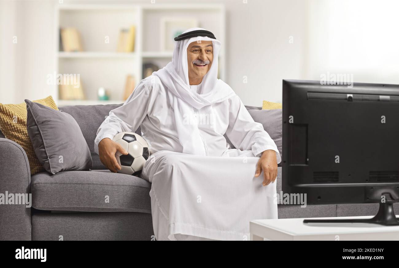 Arab man in a traditional robe holding a ball and watching football on tv at home Stock Photo