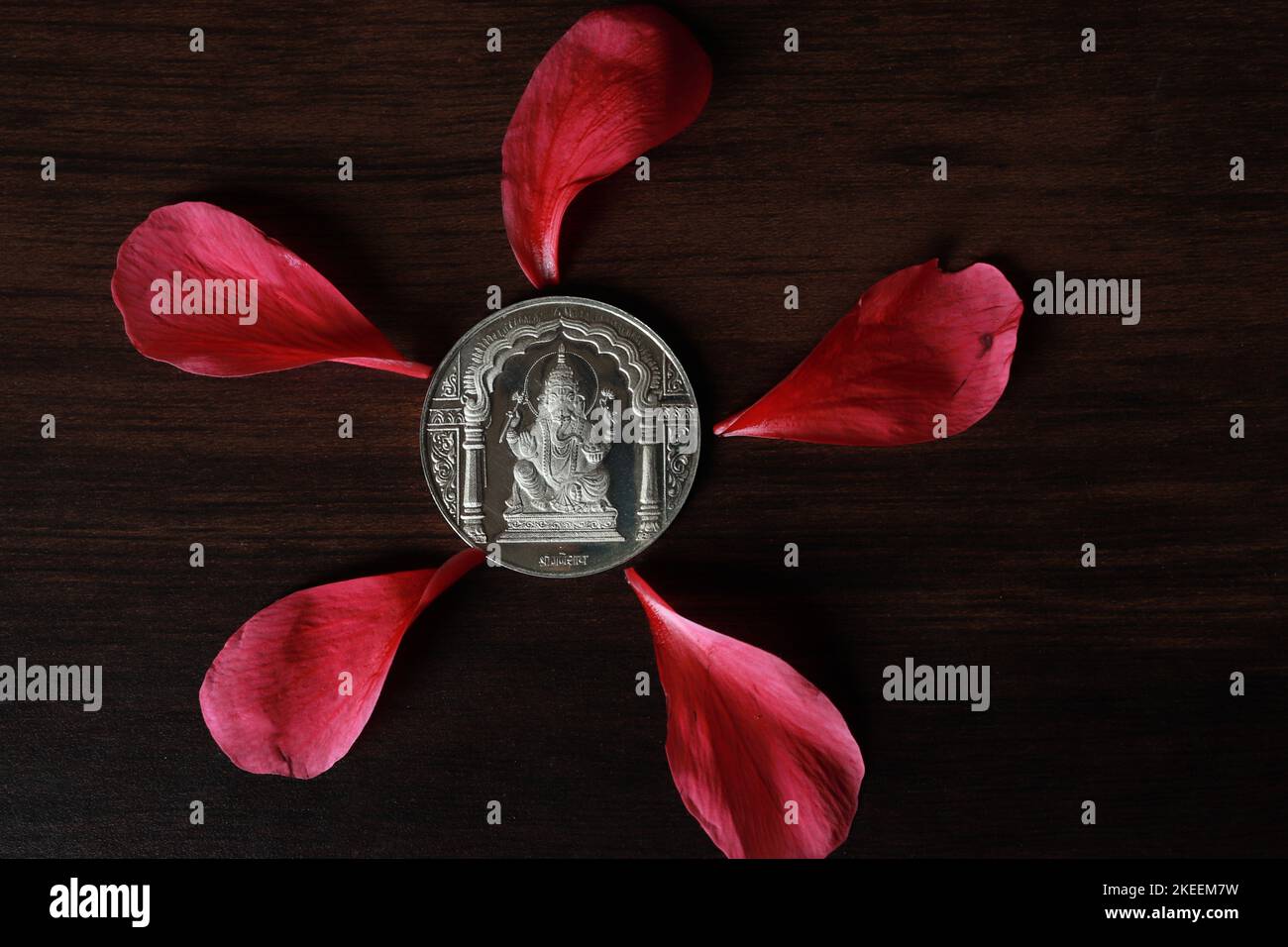 Lord Ganesha silver coin flanked by hibiscus flower petals on a wooden table/Ganesh Chaturthi Stock Photo
