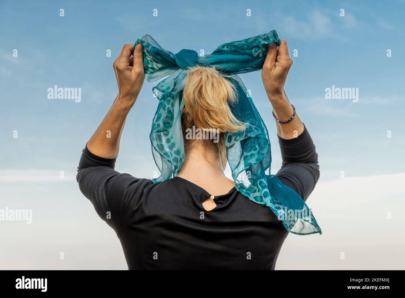 A blonde woman from behind, ties her hair with a green and blue scarf Stock Photo