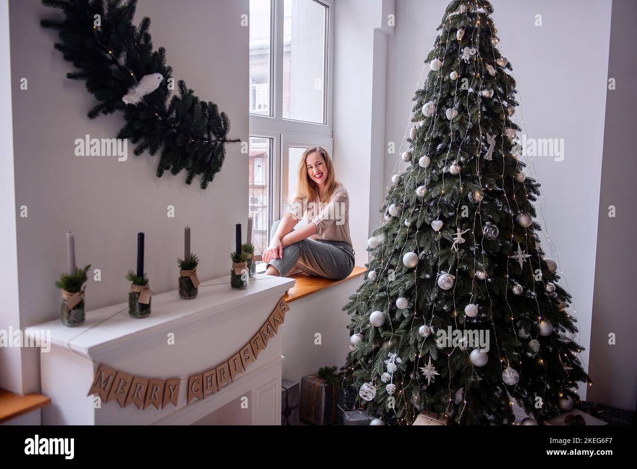 Modern stylish millennial woman sits on the windowsill near the panoramic window, decorates big Christmas tree by the fireplace, craft flags, looks ou Stock Photo