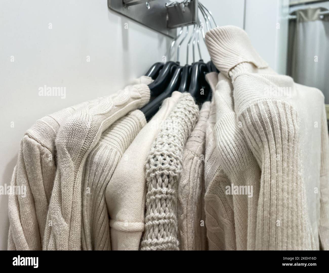 Beige color new many stack of soft cozy knitted sweaters of different design hanging on hangers in fitting room in store mall. Fall Winter season new Stock Photo