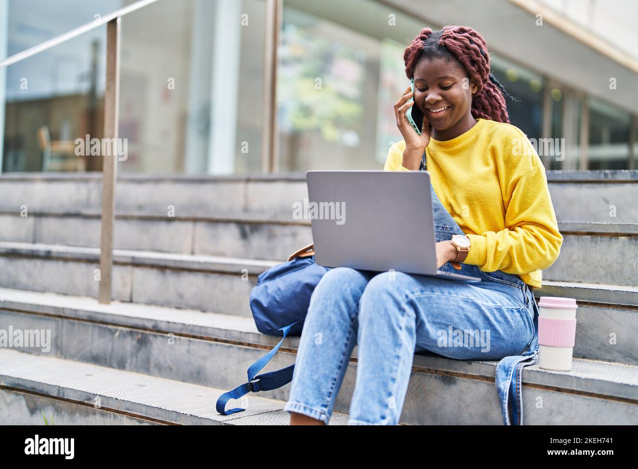African american woman student using laptop talking on smartphone at ...